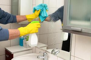 mãos do uma homem dentro amarelo luvas limpando uma espelho dentro a banheiro. foto