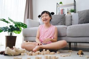 uma pequeno menina senta em a chão jogando com de madeira blocos foto