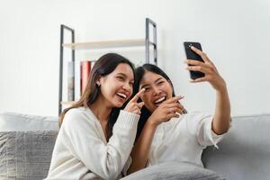 dois mulheres estão sentado em uma sofá e levando uma selfie, Veja às a inteligente telefone, e rindo. foto