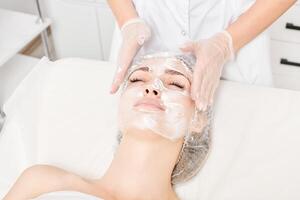 cosmetologista massageia máscara de creme na pele do rosto de mulher para rejuvenescimento, procedimento em salão de beleza foto