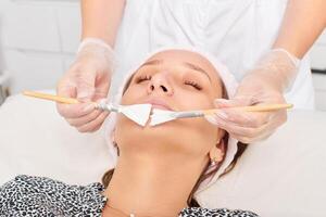 esteticista aplicando máscara de creme cosmético no rosto de mulher para rejuvenescimento, procedimento em salão de beleza foto