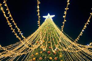 Natal árvore com amarelo guirlandas, decorativo lâmpadas e grande branco Estrela chapéu de coco às noite azul céu foto