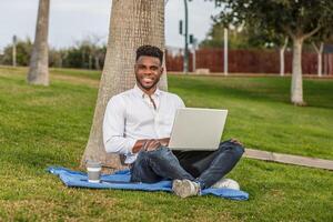 uma sorridente afro-americano homem é visto sentado em a Relva com uma computador portátil computador. foto