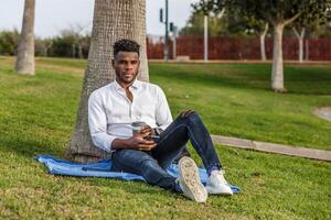 afro-americano homem sentado debaixo uma árvore usando uma célula telefone foto