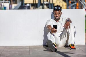 africano americano homem sentado em chão, olhando às célula telefone foto