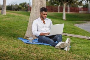 a afro-americano homem senta debaixo uma árvore, trabalhando em dele computador portátil computador. foto