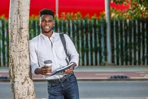 africano americano homem em pé Próximo para uma árvore com uma copo do café. foto