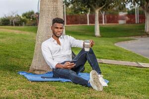 uma homem senta confortavelmente dentro a verde grama, inclinado contra uma alta árvore, segurando uma célula telefone e uma copo do café. foto