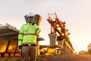 uma construção trabalhador é em pé em uma ponte, falando em dele célula telefone. a cena é conjunto dentro uma construção site, com uma ampla guindaste dentro a fundo. a trabalhador é vestindo uma amarelo Jaqueta foto