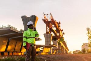 uma construção trabalhador é em pé em uma ponte, falando em dele célula telefone. a cena é conjunto dentro uma construção site, com uma ampla guindaste dentro a fundo. a trabalhador é vestindo uma amarelo Jaqueta foto