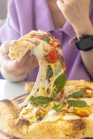 uma mulher é comendo uma pizza com dela mãos. a pizza é em uma de madeira placa. a mulher é vestindo uma roxa camisa foto