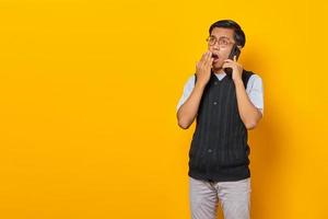jovem asiático chocado falando em um smartphone e cobrindo a boca em um fundo amarelo
