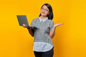 retrato de uma mulher asiática sorridente usando óculos segurando um laptop e mostrando o espaço da cópia nas palmas das mãos sobre o fundo amarelo foto