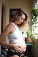 jovem grávida mulher abraçando dela grande fofa barriga enquanto em pé de a grande janela dentro vintage apartamento. a conceito do amor próprio e harmonia foto