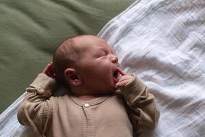 misturado raça recém-nascido bebê deitado com boca Largo aberto em verde algodão fralda vestido dentro bege maiô foto