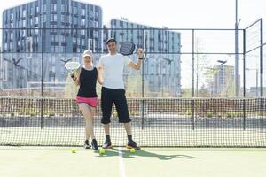 retrato do positivo jovem mulher e adulto homem em pé em padel tênis tribunal, segurando raquete e bola, sorridente foto