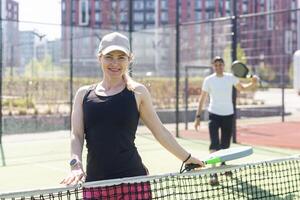 retrato do positivo jovem mulher e adulto homem em pé em padel tênis tribunal, segurando raquete e bola, sorridente foto