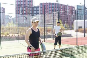 retrato do positivo jovem mulher e adulto homem em pé em padel tênis tribunal, segurando raquete e bola, sorridente foto