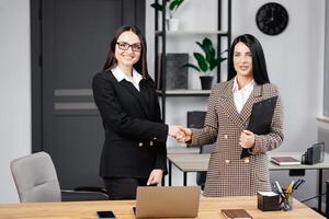 dois lindo o negócio mulheres sorridente feliz e confiante. em pé em local de trabalho e trabalhando junto, usando computador portátil e tremendo mãos para acordo às a escritório foto