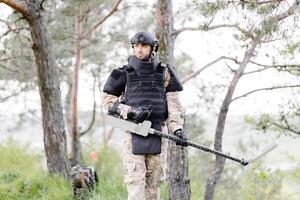 uma homem dentro uma militares uniforme e a prova de balas colete trabalho dentro a floresta com uma metal detector. uma caça-minas executa trabalhos em desminagem a território foto