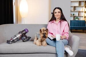 uma fofa jovem mulher é sentado em a sofá com dela cachorro e lendo uma livro e segurando uma copo dentro dela mão. foto