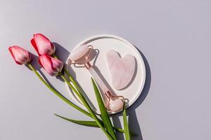rosa quartzo rolo massageador e raspador em uma volta cerâmico bandeja com viver tulipas. topo visualizar. cinzento fundo. natural Cuidado. foto