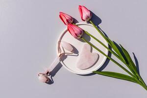 rosa quartzo rolo massageador, gua sha raspador em branco volta bandeja com viver tulipas. topo visualizar. cinzento fundo. elevação massagem, autocuidados. foto