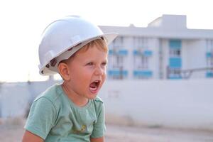 uma pequeno Garoto dentro uma branco capacete gritos. uma criança em a fundo do uma construção local. profissão - construtor. foto