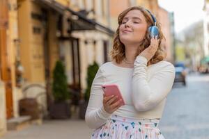 feliz jovem mulher dentro sem fio fones de ouvido escolhendo, ouvindo música dentro Smartphone dançando ao ar livre foto