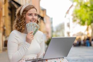 feliz rico vencedora menina usando computador portátil vitória contando dinheiro dinheiro dólar notas sentado em rua foto