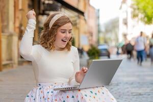 caucasiano feliz mulher trabalhando em computador portátil comemoro sucesso ganhar dinheiro sentado em urbano rua dentro cidade foto