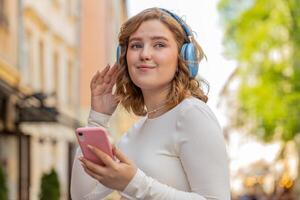 feliz jovem mulher dentro sem fio fones de ouvido escolhendo, ouvindo música dentro Smartphone dançando ao ar livre foto