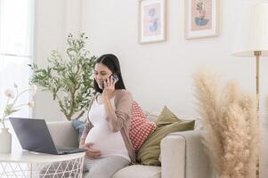 grávida mulher trabalhando em computador portátil e inteligente telefone dentro a vivo quarto às casa foto