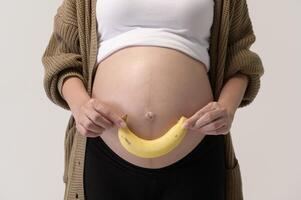 retrato do lindo grávida mulher segurando banana sobre branco fundo estúdio, saúde e maternidade conceito foto