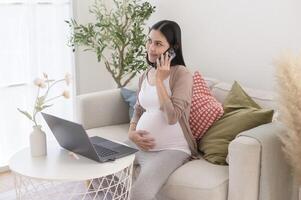 grávida mulher trabalhando em computador portátil e inteligente telefone dentro a vivo quarto às casa foto