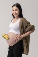 retrato do lindo grávida mulher segurando abacate sobre branco fundo estúdio, saúde e maternidade conceito foto