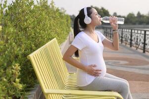 lindo grávida mulher bebendo garrafa do puro água dentro a parque, saudável e ativo gravidez estilo de vida conceito. foto