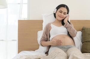 feliz grávida mulher com fones de ouvido ouvindo para Mozart música e deitado em cama, gravidez conceito foto