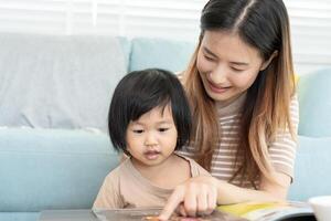 feliz mãe asiática relaxa e lê o livro com o tempo do bebê juntos em casa. pai senta no sofá com a filha e lê uma história. aprender desenvolvimento, cuidar de crianças, rir, educação, contar histórias, praticar. foto