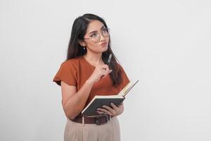 pensativo jovem ásia mulher vestindo uma Castanho camisa e Óculos segurando uma livro e olhando a parte, de lado isolado sobre branco fundo. foto