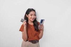 lindo jovem ásia mulher vestindo Castanho camisa é sorridente enquanto segurando dela Smartphone e crédito cartão sobre isolado branco fundo. foto