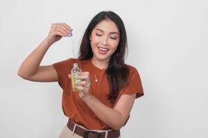 feliz jovem ásia mulher dentro Castanho camisa segurando garrafa do sérum para Aplique pele Cuidado isolado de branco fundo. foto