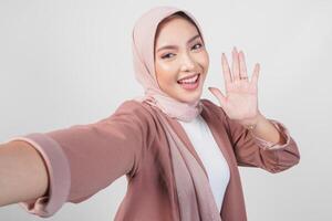 alegre jovem ásia muçulmano mulher dentro hijab posando para uma selfie com acenando mão Olá isolado sobre branco fundo. foto