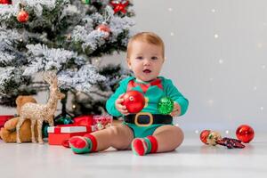 bebê dentro verde maiô detém colorida Natal bolas dentro dele mãos. criança dentro uma Natal gnomo traje foto