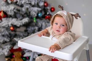 retrato do gordo bebê dentro uma macacão com veado chifres senta dentro uma branco Alto cadeira perto Natal árvore foto