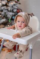 gordo bebê dentro uma macacão com veado chifres senta dentro uma branco Alto cadeira para alimentando perto Natal árvore foto