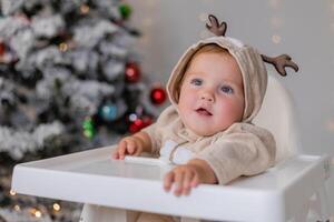 retrato do gordo bebê dentro uma macacão com veado chifres senta dentro uma branco Alto cadeira perto Natal árvore foto