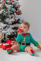 bebê dentro verde maiô detém colorida Natal bolas dentro dele mãos. criança dentro uma Natal gnomo traje foto