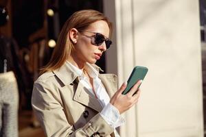 elegante europeu mulher dentro oculos de sol é olhando em telefone em cidade rua fundo foto