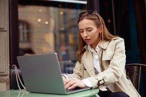 atraente europeu mulher trabalhando em computador portátil conectados enquanto sentado às ao ar livre cafeteria terraço foto